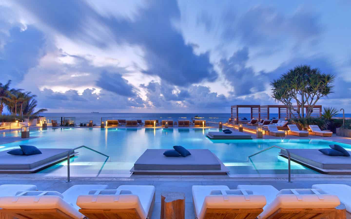 Infinity swimming pool with lounge chairs and cabanas under a cloudy blue sky at dusk